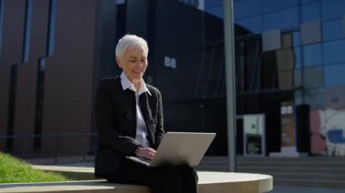 Şehir ortamında dizüstü bilgisayarla otururken videoda konuşan gülümseyen yaşlı kadın.