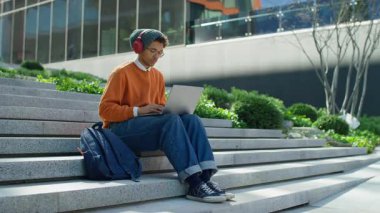 Kulaklıklı genç adam, modern binanın dışındaki merdivenlerde dizüstü bilgisayar kullanıyor, odaklanmış ve çalışıyor.