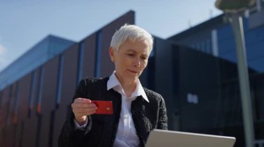 Dışarıda kredi kartı ve laptop ile online alışveriş yapan yaşlı bir kadın, şaşırmış ya da etkilenmiş görünüyor