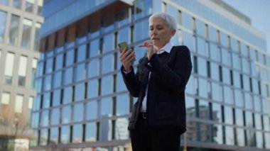 İş kıyafeti giymiş üst düzey bir kadın dışarıda akıllı telefon kullanıyor, şehir ortamında online alışveriş yaparken düşünüyor.