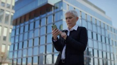 Endişeli son sınıf öğrencisi, akıllı telefondan kötü haberler okuyor, modern ofis binasının dışında başını tutuyor.