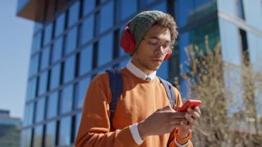 Sırt çantalı ve kulaklıklı genç adam dışarıda cam binanın önünde yürürken telefonla mesajlaşıyor.