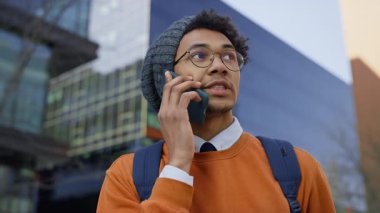 Sırt çantalı ve bereli genç bir öğrenci cam üniversite binasının yanında telefonla konuşuyor, ciddi görünüyor.