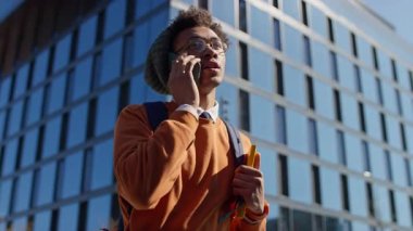 Sırt çantalı ve bereli bir adam telefonda konuşuyor, elinde defter, modern ofis binasının yanında duruyor.