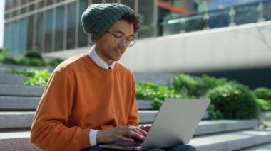 Genç adam dışarıda dizüstü bilgisayarda kompozisyon yazarken gülümsüyor, üniversite binasının yanındaki merdivenlerde oturuyor.