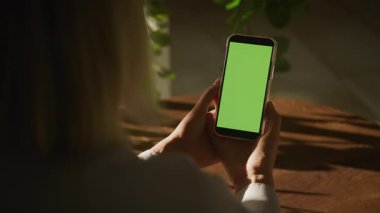 Evinde yeşil ekranlı akıllı telefon kullanan bir kadının yakın plan fotoğrafı. Doğal ışık ve kapalı alan bitkileri var..