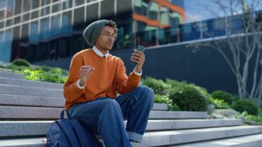 Genç adam modern binanın yanındaki merdivenlerde oturuyor, telefonda konuşurken el kol hareketi yapıyor, rahat kolej ortamında.