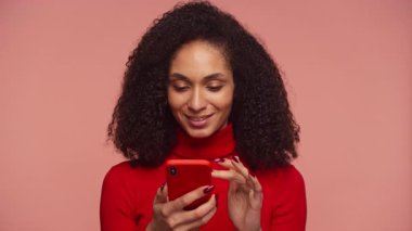 Çevrimiçi alışveriş yapmak için akıllı telefonu kullanan mutlu bir kadın. Stüdyoda pembe arka planda kırmızı balıkçı yakalı kazak giyiyor.