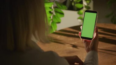 Kapalı bir ortamda yeşil ekranlı akıllı bir telefondan çalan kişi, arka planda güneş ışığı ve ev bitkileri..