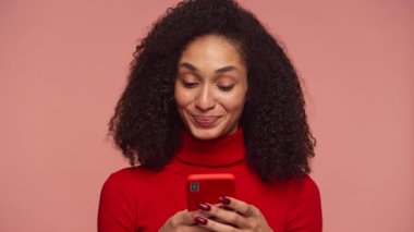 Mesajlaşırken akıllı telefona bakan neşeli bir kadın. Stüdyoda pembe arka planı olan kırmızı balıkçı yaka bir kazak giyiyor.