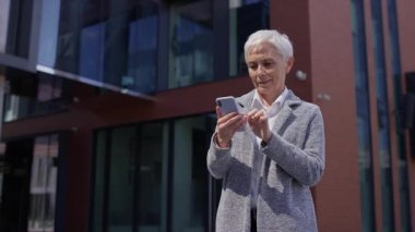 Olgun bir kadın dışarıda akıllı telefon kullanıyor, modern ofis binalarının yakınında kaydırıyor ve okuyor.