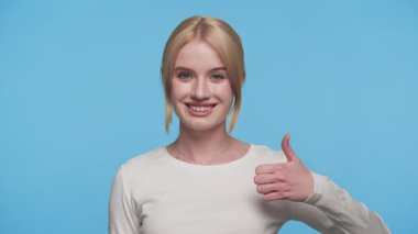 Gülümseyen ve onay jestinde temiz mavi arka plana karşı baş parmak gösteren olumlu genç kadın.