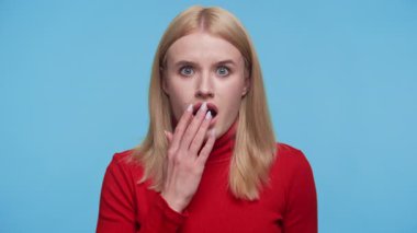 Kırmızı balıkçı yakalı genç kadın ağzını geniş gözlerle kapatıyor. Mavi arka plandaki beklenmedik haberlere tepki veriyor..