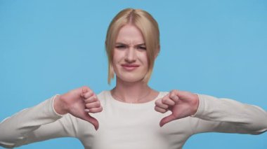 Mutsuz, beyaz bluzlu genç bir kadın hayal kırıklığına uğramış bir yüz ifadesiyle mavi arka planda baş parmak hareketi yapıyor..