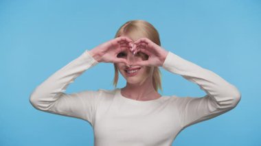 Beyazlar içinde neşeli genç bir kadın. Elleri gözlerinin üzerinde kalp şekli oluşturuyor. Sevgi ve neşe ifade ediyor..