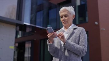Neşeli olgun bir kadın dışarıda akıllı telefona bakarken tezahürat yapıyor, bir iş veya kişisel bağlamda heyecan ve başarı ifade ediyor..