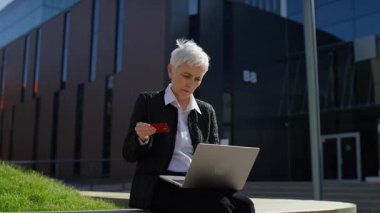 Modern bir ofis binasının önünde dizüstü bilgisayar kullanırken elinde kredi kartı tutan yaşlı bir kadın e-ticaret ve finans sektörünü temsil ediyor..