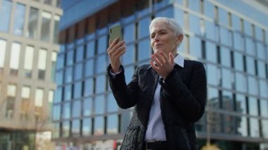 Mutlu yaşlı bir kadın, modern bir ofis binasının dışındaki akıllı telefonuyla iletişim ve bağlantı kurduğunu belirten bir video görüşmesi yapıyor..