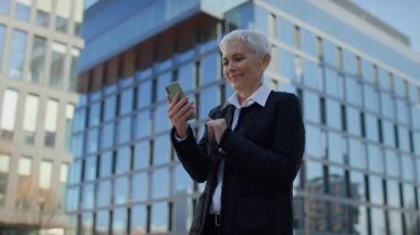 Neşeli yaşlı kadın, akıllı telefonundan iyi haberler okurken, modern cam ofis binasının önünde dururken olumlu tepki veriyor..