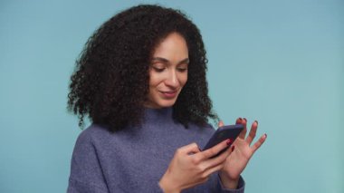 Kıvırcık saçlı neşeli genç kadın akıllı telefonuna bakarken seviniyor, iyi haberi ya da başarıyı kutluyor, mavi arka planda izole edilmiş..