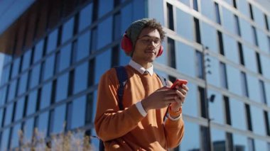 Kulaklık ve bere takan modaya uygun genç bir adam modern cam ofis binasının önünde akıllı telefonunu kullanırken müzik dinlemekten hoşlanıyor..