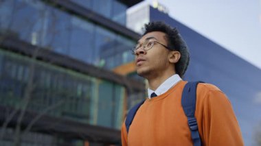 Sırt çantası ve günlük giysiler giyen kararlı genç bir öğrenci modern bir şehir bölgesinde yürüyor, eğitime gidiyor ya da derse katılıyor..