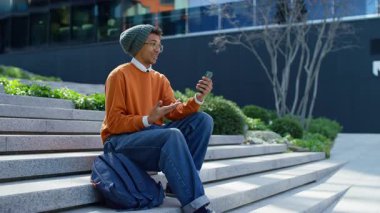 Sırt çantasıyla merdivenlerde oturan neşeli genç bir öğrenci, bir şehir kampüsünde akıllı telefonuyla video görüşmesi yaparken heyecanla konuşuyor..