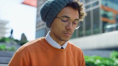 Bere takan ve gözlük takan genç bir adam modern bir üniversite binasının önünde merdivenlerde otururken dizüstü bilgisayarda dikkatle çalışıyor..