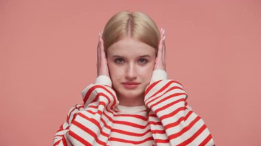 Çizgili gömlekli genç bir kadın, kulaklarını elleriyle kapatıyor, hayal kırıklığını ifade ediyor ya da pembe arka planda dinlemeyi reddediyor..