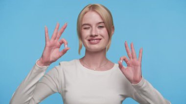 Gülümseyen ve göz kırpan neşeli genç bir kadın mavi arka plana karşı iki kere tamam el hareketi yapıyor.
