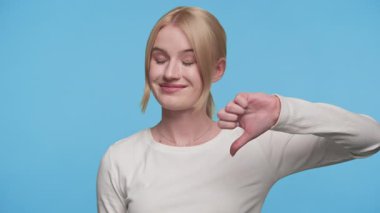 Sarışın kadın onaylamayan yüzüyle başparmak kaldırıyor. Stüdyoda mavi arka planda izole edilmiş.
