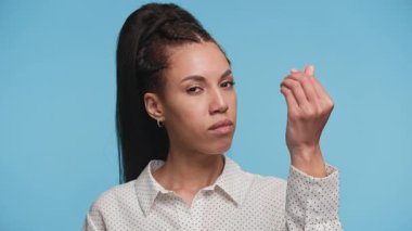 Kendine güvenen genç bir kadın el hareketi yapıp para talep ediyor, mavi arka planda duruyor ve beyaz benekli tişört giyiyor..