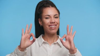 Kendinden emin genç bir kadın gülümserken mavi arka planda çift onay işareti yapıyor, pozitifliğini ve onayını ifade ediyor..