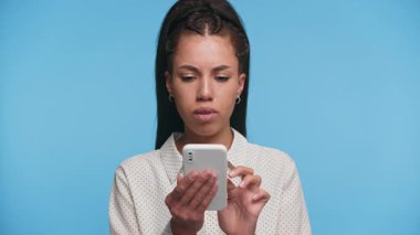 Endişeli genç kadın, mavi arka planda stres ve güvensizlik gösteren, akıllı telefondan rahatsız edici bir mesaj okuyor..