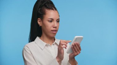 Gülümseyen genç kadın akıllı telefonunda kayar, içerikten zevk alır ya da parlak mavi bir arkaplanda uygulamaları tarar.