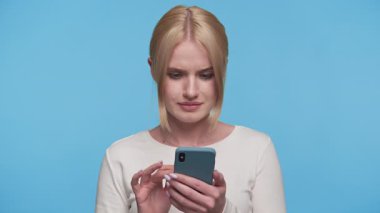 Genç bir kadın üzgün ve kafası karışmış bir halde elinde akıllı telefonla mavi arka plandaki şaşırtıcı ve nahoş bir mesaja tepki veriyor..