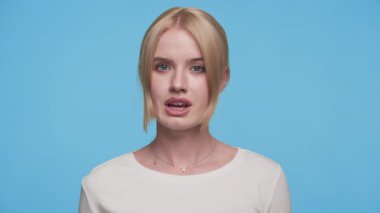 Parmakları dudaklarına değen genç bir kadın sessizlik ya da gizlilik işareti veriyor, mavi bir arka plana karşı duruyor ve ciddi görünüyor..