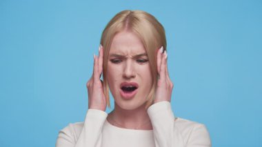Genç bir kadın rahatsız bir şekilde şakaklarına ve buruşukluklarına dokunuyor. Mavi arka planda baş ağrısı ya da stres belirtileri gösteriyor..