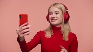 Kırmızı kulaklık takan neşeli genç bir kadın bir video konuşmasında pembe arka plana karşı akıllı telefonunu kullanarak gülümsüyor..