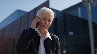 Modern bir ofis binasının önünde dizüstü bilgisayarla konuşurken profesyonel kıyafetli yaşlı bir kadın telefonda konuşuyor..