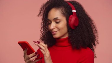 Kırmızı kulaklık takan neşeli bir kadın ve balıkçı yaka kazak. Akıllı telefonunu kaydırıyor ve ekranda olumlu bir şeyler okurken gülüyor..