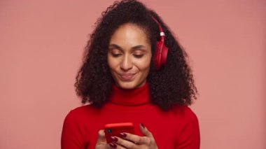 Kırmızılı genç kadın, kulaklıkla müzik dinlerken ve elinde akıllı telefon tutarken huzur içinde gülümsüyor..
