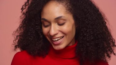 Kıvırcık saçlı, pembe arka planda kırmızı balıkçı yaka kazak giyen genç bir kadının neşeli yakın çekimi..