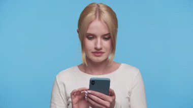 Şok olmuş genç bir kadın, parlak mavi bir arka planda akıllı telefonuna bakarken ağzını inançsızlıkla kapatıyor..