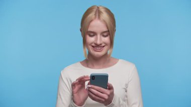 Gülümseyen genç kadın, mavi arka planda pozitif bir çevrimiçi alışveriş ya da gezinme deneyimi önererek akıllı telefonu neşeyle kullanır..