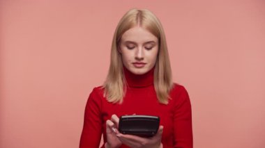 Kırmızı balıkçı yakalı genç sarışın kadın hesap makinesi kullanırken odaklanıyor, muhtemelen pembe arka planda bütçe veya finansal planlama yapıyor..