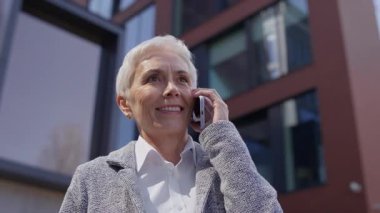 Beyaz saçlı kendinden emin yaşlı kadın telefonda konuşurken gülümsüyor modern şehir ortamında iletişim ve başarı gösteriyor..