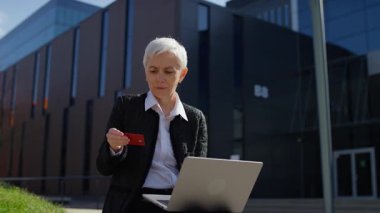 Dışarıdaki yaşlı iş kadını kredi kartına endişeli bir şekilde bakıyor, cep telefonu ödemeleri sırasında yapılan işlemin başarısız olduğunu veya geri çevrildiğini gösteriyor.