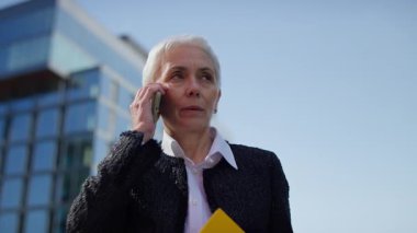 Kendinden emin yaşlı bir kadın, modern bir ofis binasının dışındaki telefon görüşmesinde konuşurken sarı bir klasör tutuyor..