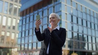 Akıllı telefonuyla yaptığı bir video görüşmesi sırasında gülümseyen yaşlı kadın modern bir şehrin iş bölgesinde dışarıda duruyor..